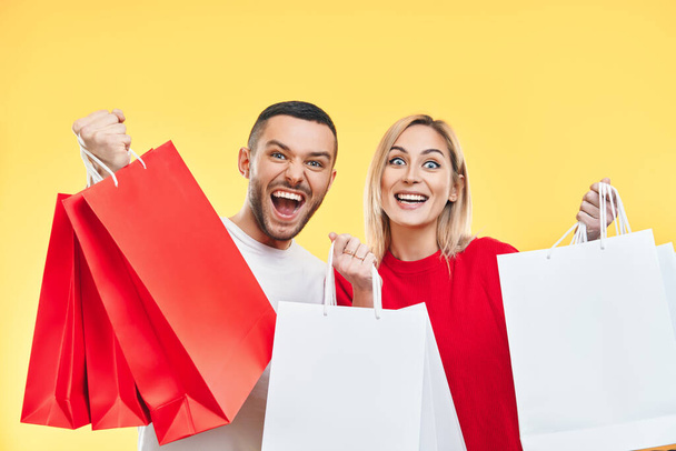 Young happy couple holding shopping bags in hands isolated over yellow background. shopping, sale, shopaholic concept - Fotoğraf, Görsel