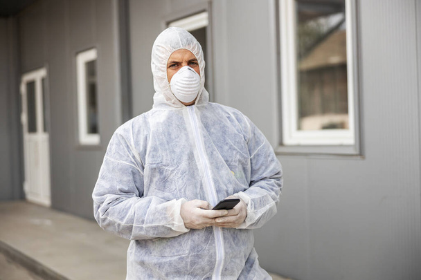 Virüs koruyucu giysili ve maskeli bir adam cep telefonu akıllı telefonuyla bakıyor ve daktilo ediyor. Koronavirüs binalarını spreyle dezenfekte ediyor. Enfeksiyon önleme ve salgının kontrolü. Dünya salgını.  - Fotoğraf, Görsel