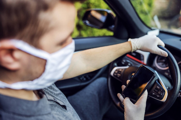 Koruyucu maskeli ve eldivenli bir adam elinde cep telefonu olan bir araba kullanıyor. Enfeksiyon önleme ve salgının kontrolü. Dünya salgını. Kendine dikkat et.. - Fotoğraf, Görsel