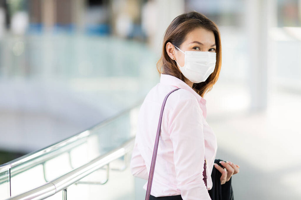 Beautiful adult middle age Asian working office businesswoman wearing surgical protective medical mask on face looking straight to camera while walking in path way to work in office. Health care concept. - Photo, Image