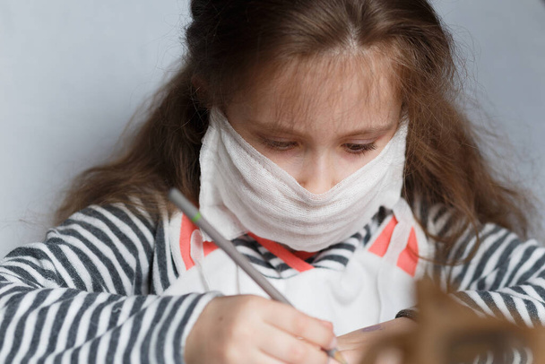 ute weinig Kaukasisch kind meisje dragen van een beschermende masker en tekent in potlood aan een tafel in een kinderkamer - Foto, afbeelding