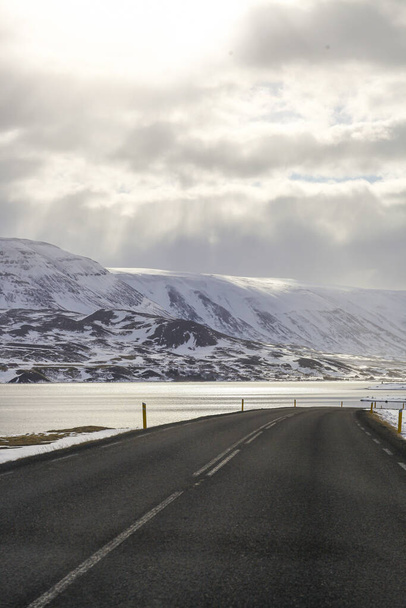 Σκοτεινός ασφαλτοστρωμένος δρόμος στο φόντο των βουνών και του ωκεανού. Η περιοδεία στην Ισλανδία. Γεωθερμική θερμότητα από εδώ. - Φωτογραφία, εικόνα