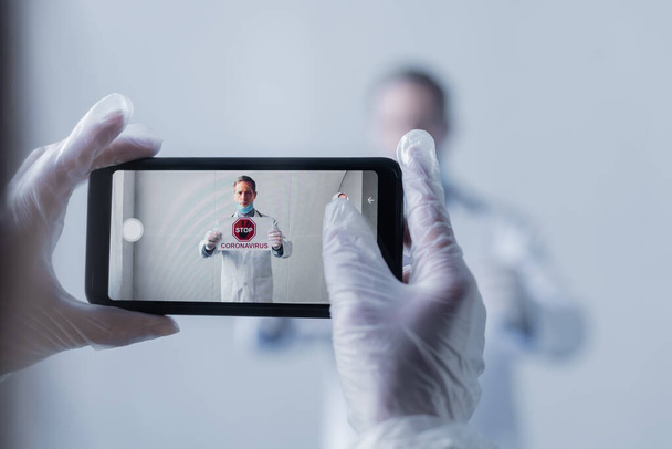 selectieve focus van arts het nemen van foto van volwassen man in witte jas met bordje stop coronavirus belettering op grijs - Foto, afbeelding