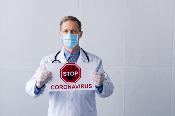 Врач средних лет в белом халате и медицинской маске держит плакат с остановкой коронавируса на сером
 - Фото, изображение