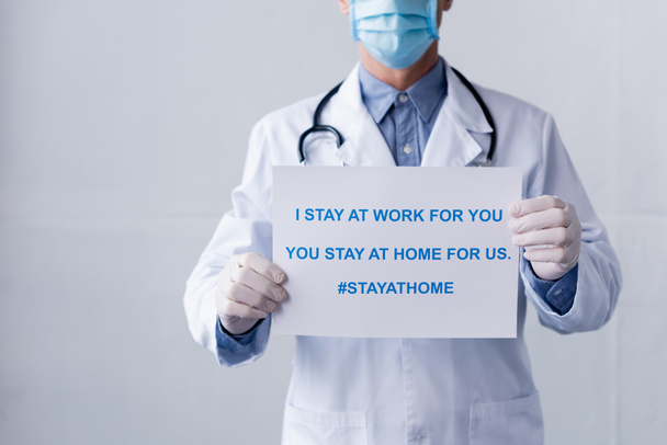 cropped view of middle aged doctor in latex gloves holding placard with i stay at work for you, you stay at home for us lettering on grey - Фото, зображення