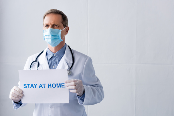 middle aged doctor in latex gloves holding placard with stay at home lettering on white  - Foto, Bild