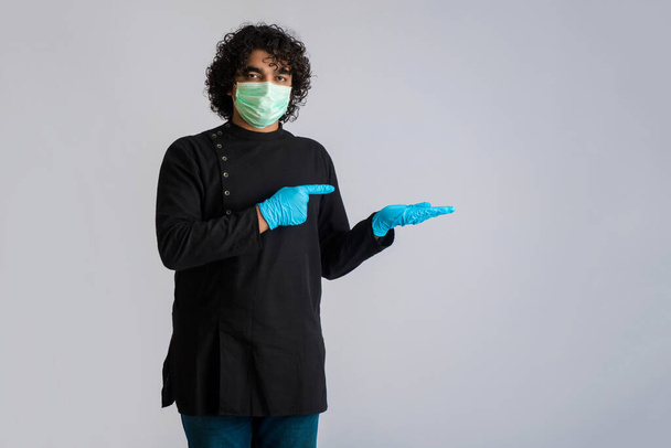 Молодой человек в медицинской маске показывает что-то под рукой. мужчина в хирургической маске для защиты от коронавируса
. - Фото, изображение