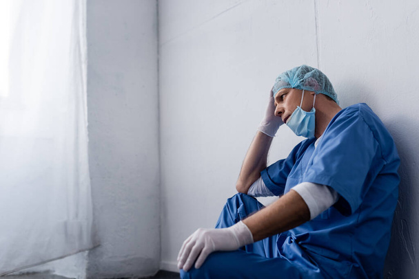 medico stanco e maturo in berretto medico e occhiali seduti vicino al muro bianco in clinica
 - Foto, immagini