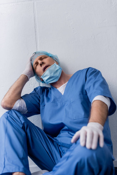 unavený a zralý lékař se zavřenýma očima sedí u bílé stěny - Fotografie, Obrázek