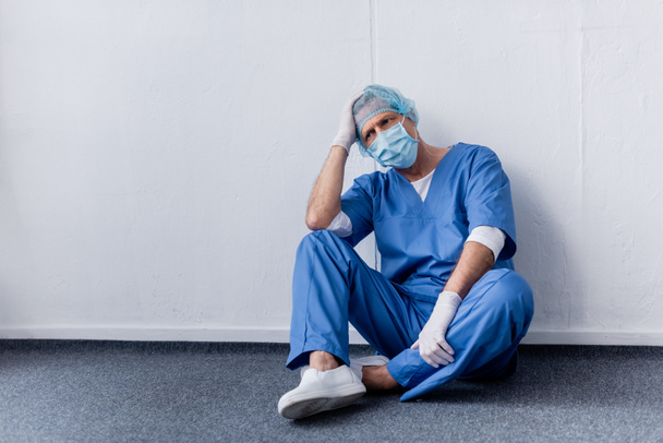 médico cansado y de mediana edad en gorra médica y gafas sentado cerca de la pared blanca
 - Foto, Imagen