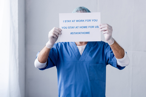mature doctor covering face with placard with i stay at work for you, you stay at home for us lettering on white  - Foto, Bild