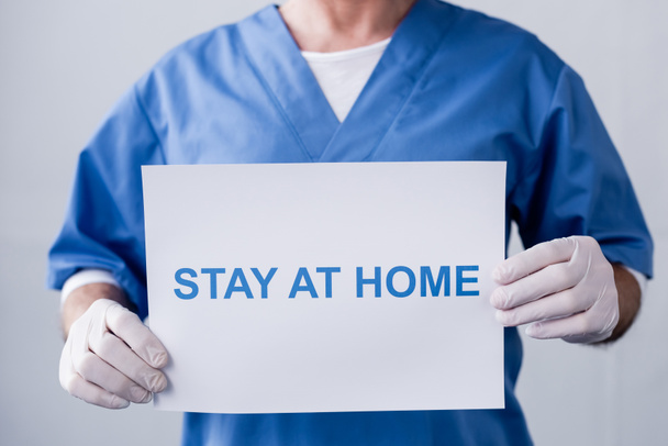 cropped view of mature doctor holding placard with stay at home lettering on grey - 写真・画像