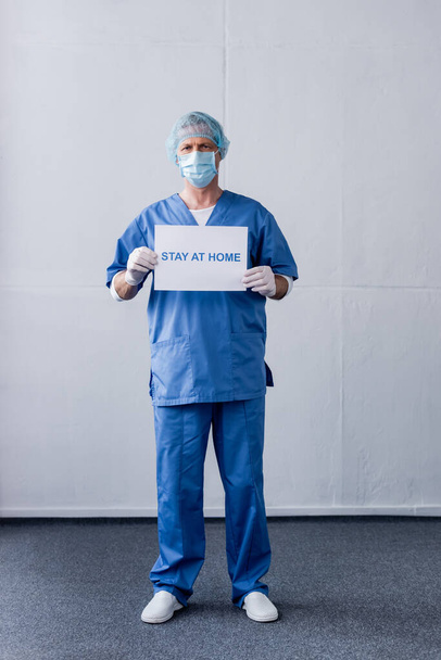 mature doctor in medical mask, cap and latex gloves holding placard with stay at home lettering while standing on grey - Фото, изображение