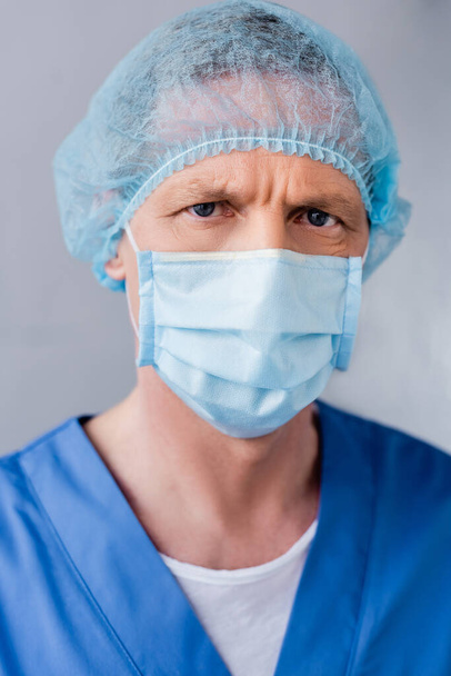 Mavi maskeli ve şapkalı olgun bir doktor grideki kameraya bakıyor.  - Fotoğraf, Görsel
