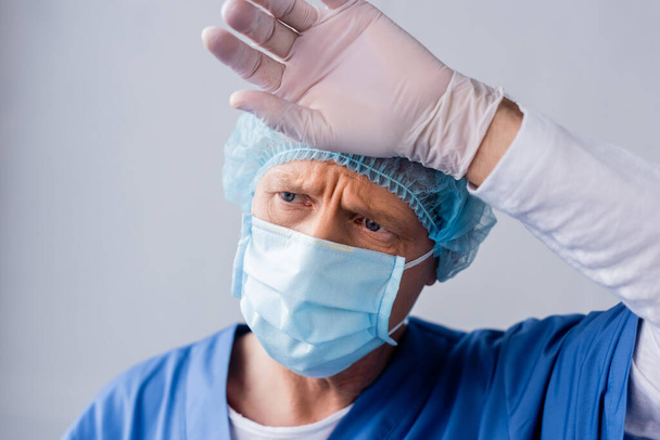 médico cansado y maduro en la máscara médica azul y la tapa tocando la frente en gris
  - Foto, Imagen