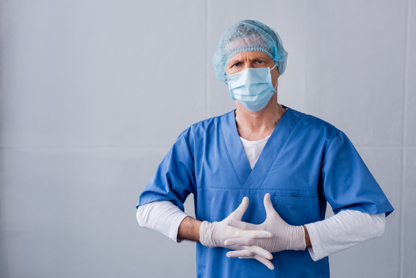 médico maduro en máscara médica azul, gorra y guantes de látex con las manos cerradas en gris
 - Foto, Imagen