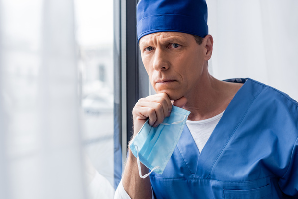 σκεπτικός και ώριμος γιατρός σε μπλε καπέλο scrub κρατώντας ιατρική μάσκα και κοιτάζοντας την κάμερα  - Φωτογραφία, εικόνα