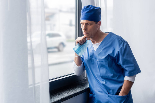 médico pensativo y maduro en sombrero de exfoliante azul que sostiene la máscara médica, de pie con la mano en el bolsillo y mirando a la ventana
  - Foto, Imagen