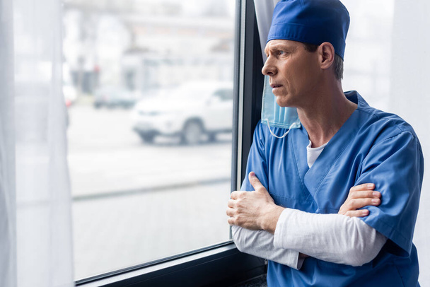 Dojrzały lekarz w niebieskim kapeluszu i masce medycznej, stojąc z skrzyżowanymi ramionami i patrząc na okno  - Zdjęcie, obraz