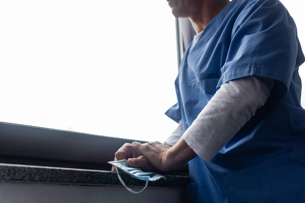 przycięty widok dojrzałego lekarza trzymającego maskę medyczną i stojącego w pobliżu okna w szpitalu  - Zdjęcie, obraz