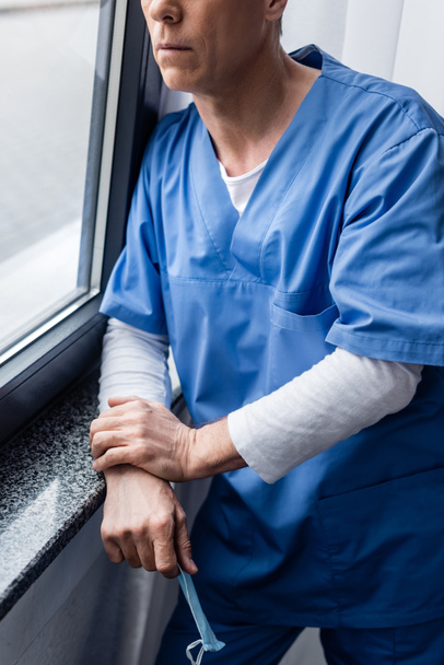 Ausgeschnittene Ansicht eines Arztes mittleren Alters mit medizinischer Maske, der am Fenster im Krankenhaus steht  - Foto, Bild