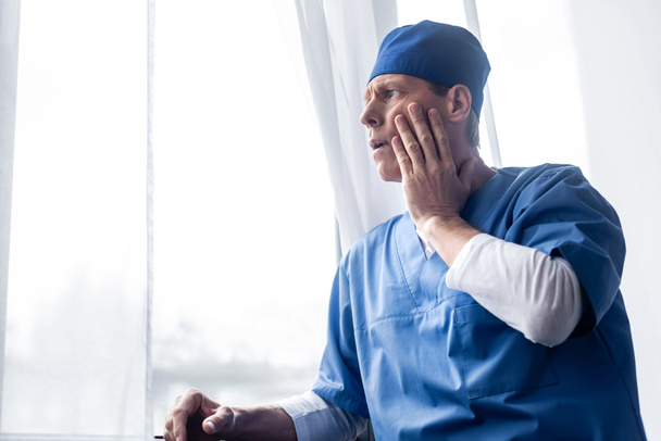 médico cansado e de meia-idade em chapéu de esfoliação de pé perto da janela no hospital e rosto comovente
 - Foto, Imagem