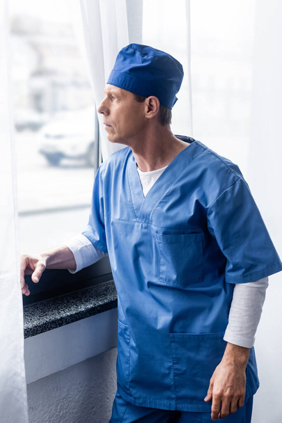 Arzt mittleren Alters mit blauem Schrubberhut blickt auf Fenster  - Foto, Bild