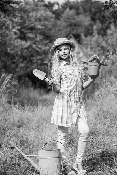Child in hat with shoulder blade small shovel hoe. Happy smiling gardener girl. Ranch girl. Happy childhood. Planting plants. Little kid hold flower pot. Spring country works. Happy childrens day - Фото, изображение