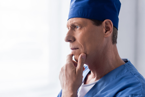 pensive middle aged doctor in scrub hat touching face while thinking on white - Photo, Image