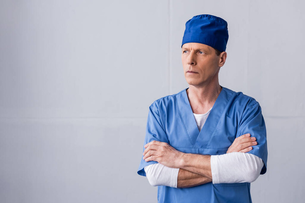 médico de mediana edad pensativo en sombrero de exfoliante azul de pie con los brazos cruzados en gris
 - Foto, Imagen