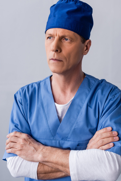 médico pensativo y maduro en sombrero de exfoliante azul de pie con los brazos cruzados en gris
 - Foto, Imagen