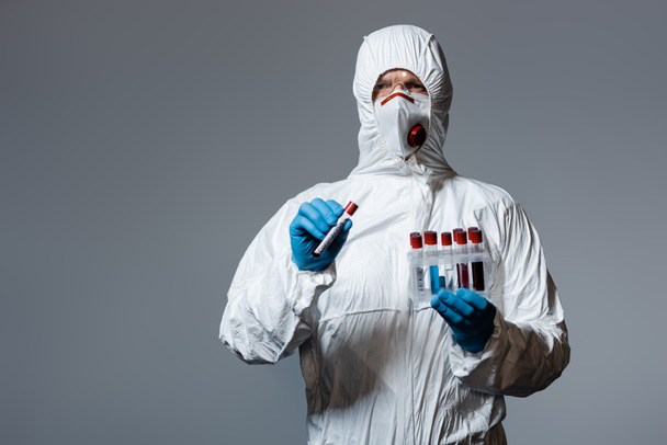 ハズマットスーツの成人医師医療用マスクとゴーグル灰色に隔離されたコロナウイルスの文字でテスト管を保持  - 写真・画像
