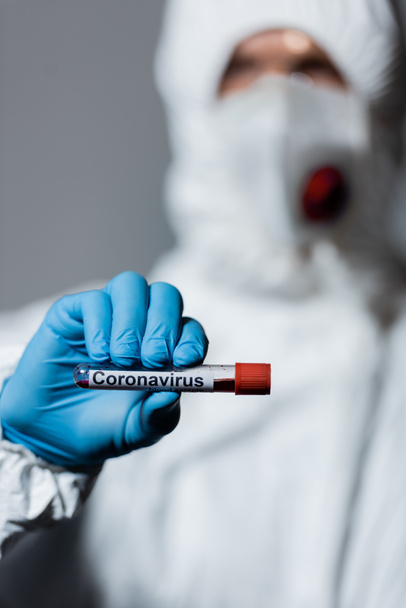 selective focus of mature doctor in hazmat suit, medical mask and goggles holding test tube with coronavirus lettering isolated on grey  - Photo, Image