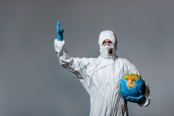 зрелый врач в защитном костюме, медицинской маске и латексных перчатках, держащих глобус и жестикулирующих изолированно на сером
  - Фото, изображение