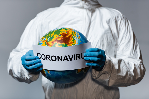 cropped view of man in hazmat suit holding globe and paper with coronavirus lettering isolated on grey  - Photo, Image