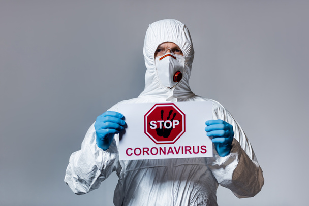 зрелый врач в защитном костюме с плакатом с остановкой коронавируса, изолированным на сером
 - Фото, изображение