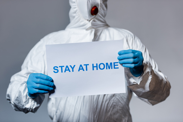 cropped view of man in hazmat suit holding placard with stay at home lettering isolated on grey - Foto, immagini
