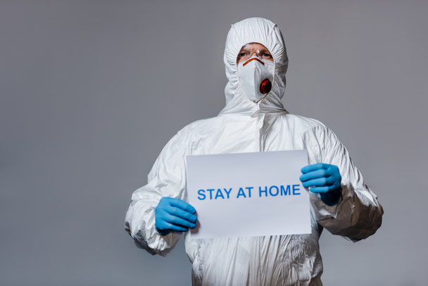 mature doctor in hazmat suit holding placard with stay at home lettering isolated on grey - Fotografie, Obrázek