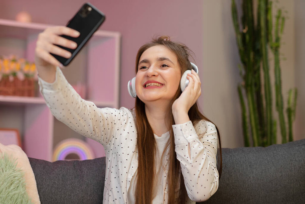 Beautiful woman listening to music at home. Pretty smiling girl taking selfie on smartphone, listening to music, sitting on couch at home. - Fotoğraf, Görsel