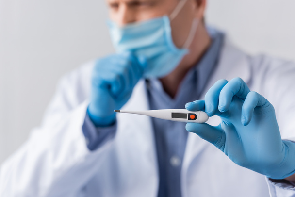 selective focus of mature doctor in latex gloves holding digital thermometer and coughing isolated on grey - Foto, Imagen