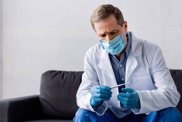 volwassen arts in medisch masker en latex handschoenen kijken naar digitale thermometer terwijl zitten op grijs - Foto, afbeelding