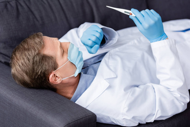 зрілий лікар в медичній масці і латексних рукавичках дивиться на цифровий термометр, лежачи і кашляючи на дивані
 - Фото, зображення