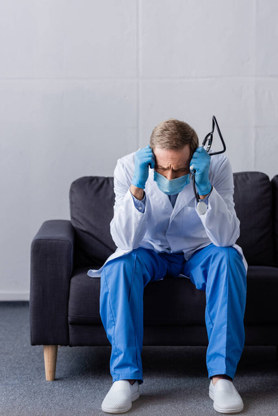 fáradt érett orvos migrén gazdaság sztetoszkóp, miközben ül a kanapén  - Fotó, kép