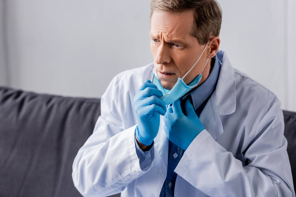 расстроен зрелый доктор в латексных перчатках касаясь медицинской маски
  - Фото, изображение