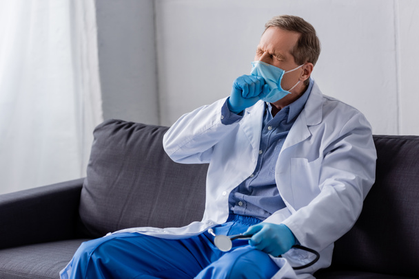 médico cansado y maduro en máscara médica y guantes de látex sentado en el sofá
  - Foto, Imagen