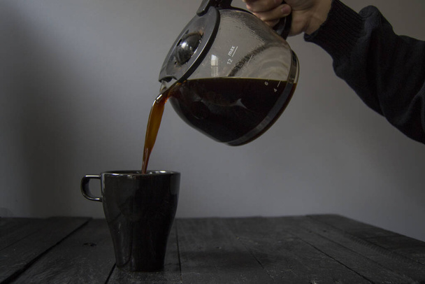 Glass jug pouring coffee into a black mug set on black wood and gray background. - Photo, Image