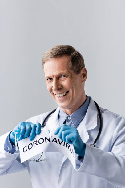 fröhlicher und reifer Arzt in Latexhandschuhen, der Papier mit Coronavirus-Schriftzug isoliert auf grau reißt - Foto, Bild