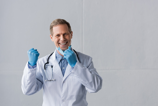 szczęśliwy i dojrzały lekarz w lateksowych rękawiczkach i masce medycznej z zaciśniętej pięści na szarości - Zdjęcie, obraz