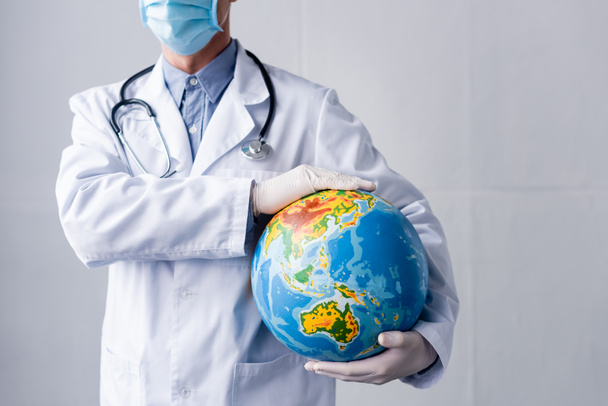 przycięty widok dojrzałego lekarza w lateksowych rękawiczkach i maskach medycznych trzymających kulę ziemską na szarości  - Zdjęcie, obraz