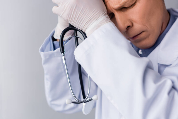 uitgeput en volwassen arts met gesloten ogen houden stethoscoop geïsoleerd op grijs - Foto, afbeelding